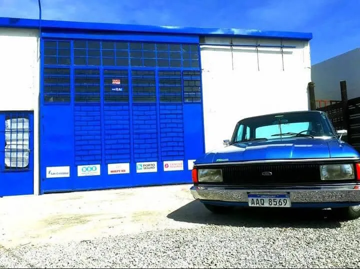 Fachada azul del Taller Brenma en Uruguay, con las calcomanías de las compañías de seguros y un Ford Falcon estacionado al frente